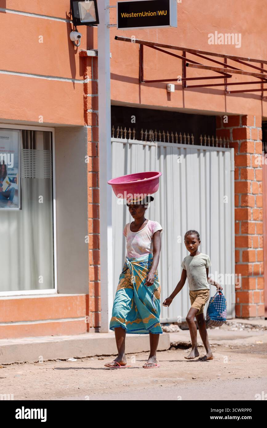 Miandrivazo, Madagascar - 11 ottobre 2025: Scena della vita quotidiana a Miandrivazo, dove una donna trasporta merci sulla testa vicino a un'azienda moderna. Foto Stock