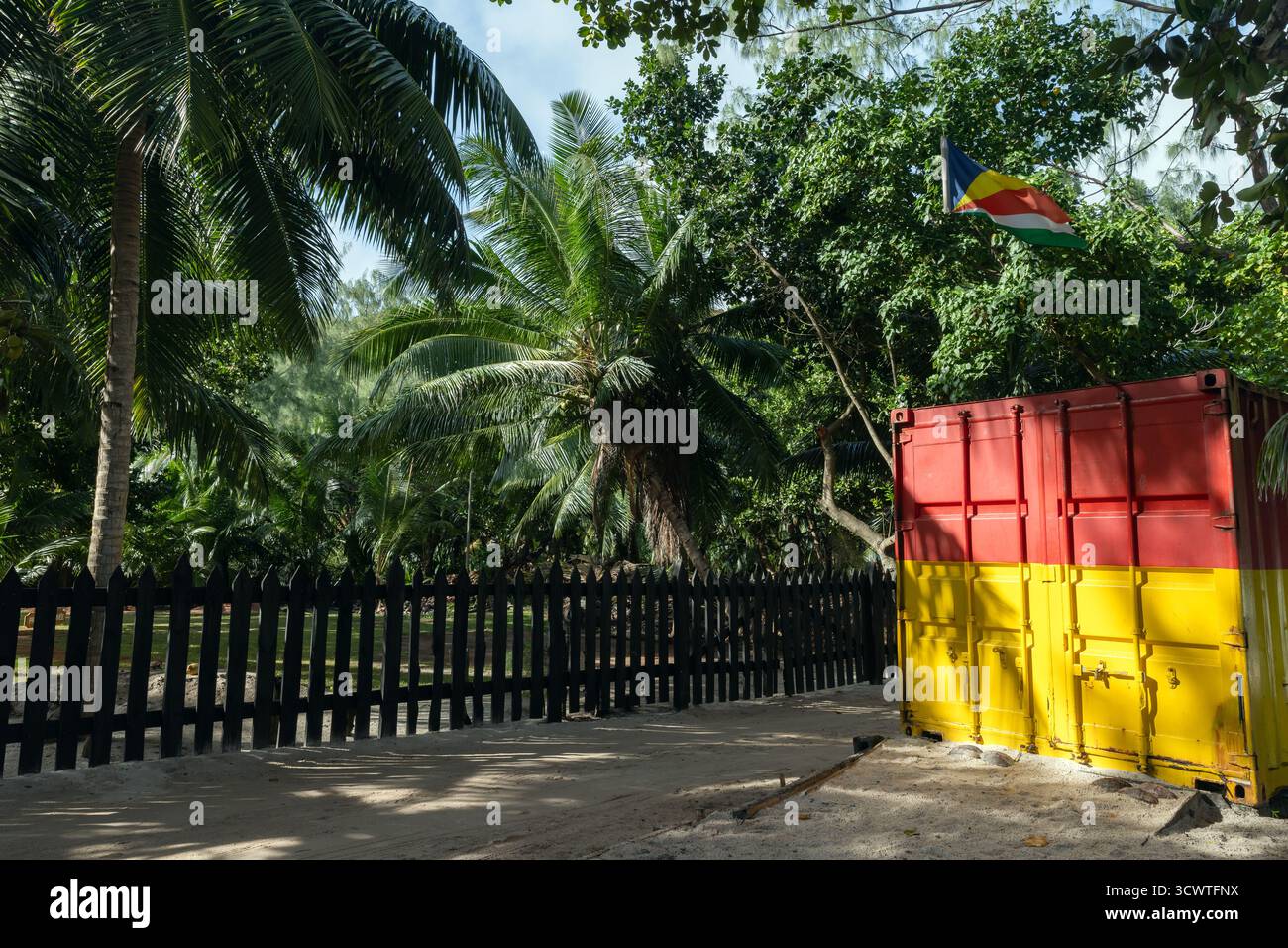 Una soleggiata scena tropicale con alte palme, una recinzione di legno scuro e un contenitore di spedizione rosso-giallo luminoso. Una bandiera sventola sopra la testa. Seychelles, Pr Foto Stock