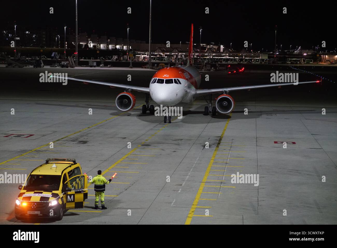 Airbus A320 easyJet guidato da un marshaler e un veicolo follow-me di notte sul grembiule dell'aeroporto di Monaco sotto i riflettori. Monaco, Germania Foto Stock