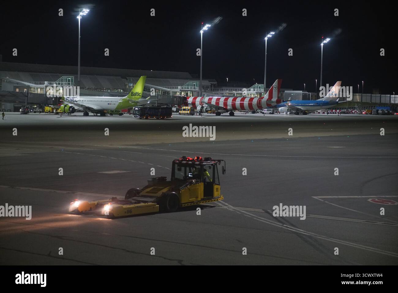Trattore che si sposta di notte sul grembiule dell'aeroporto di Monaco, con aeromobili parcheggiati e terminal illuminato sullo sfondo.Monaco, Germania Foto Stock