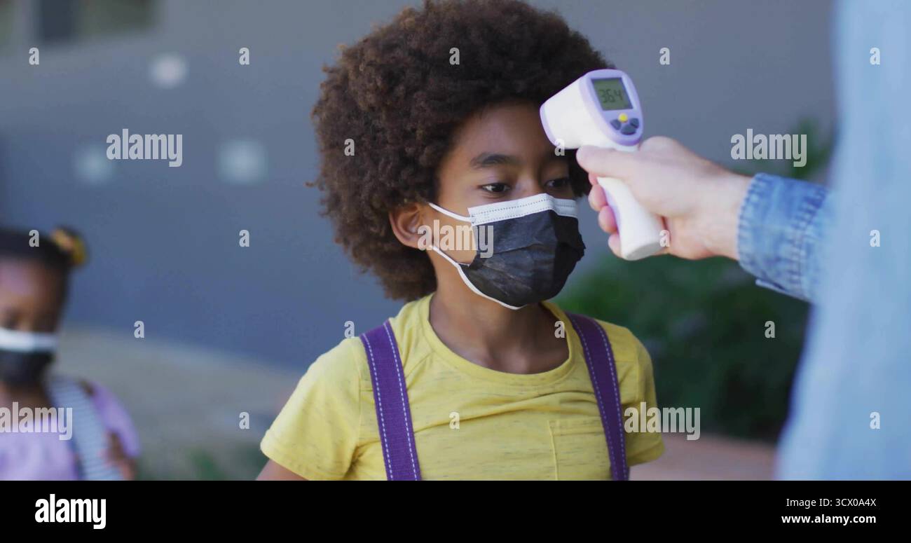 Tenendo in mano il termometro digitale, il personale controlla la fronte dello studente all'ingresso, con zaini e maschere Foto Stock