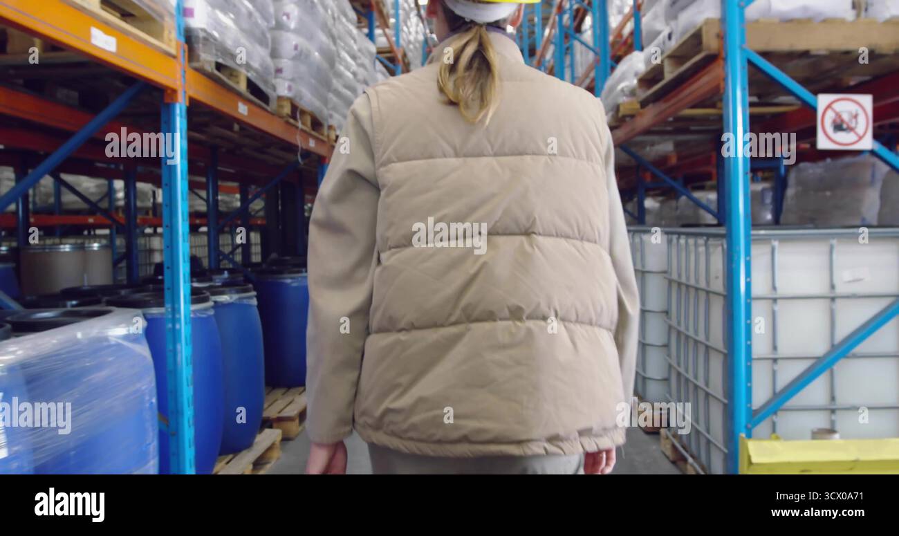 Lavoratore di magazzino che cammina indossando elmetto bianco, giubbotto abbronzante tra alti scaffali in acciaio, con tamburi blu Foto Stock