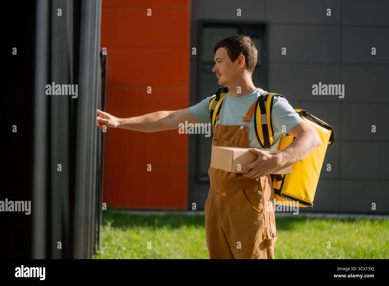 Giovane che lavora come corriere, indossa tute marroni e una borsa termica gialla, consegna un piccolo pacco all'indirizzo di un cliente e chiama il campanello Foto Stock