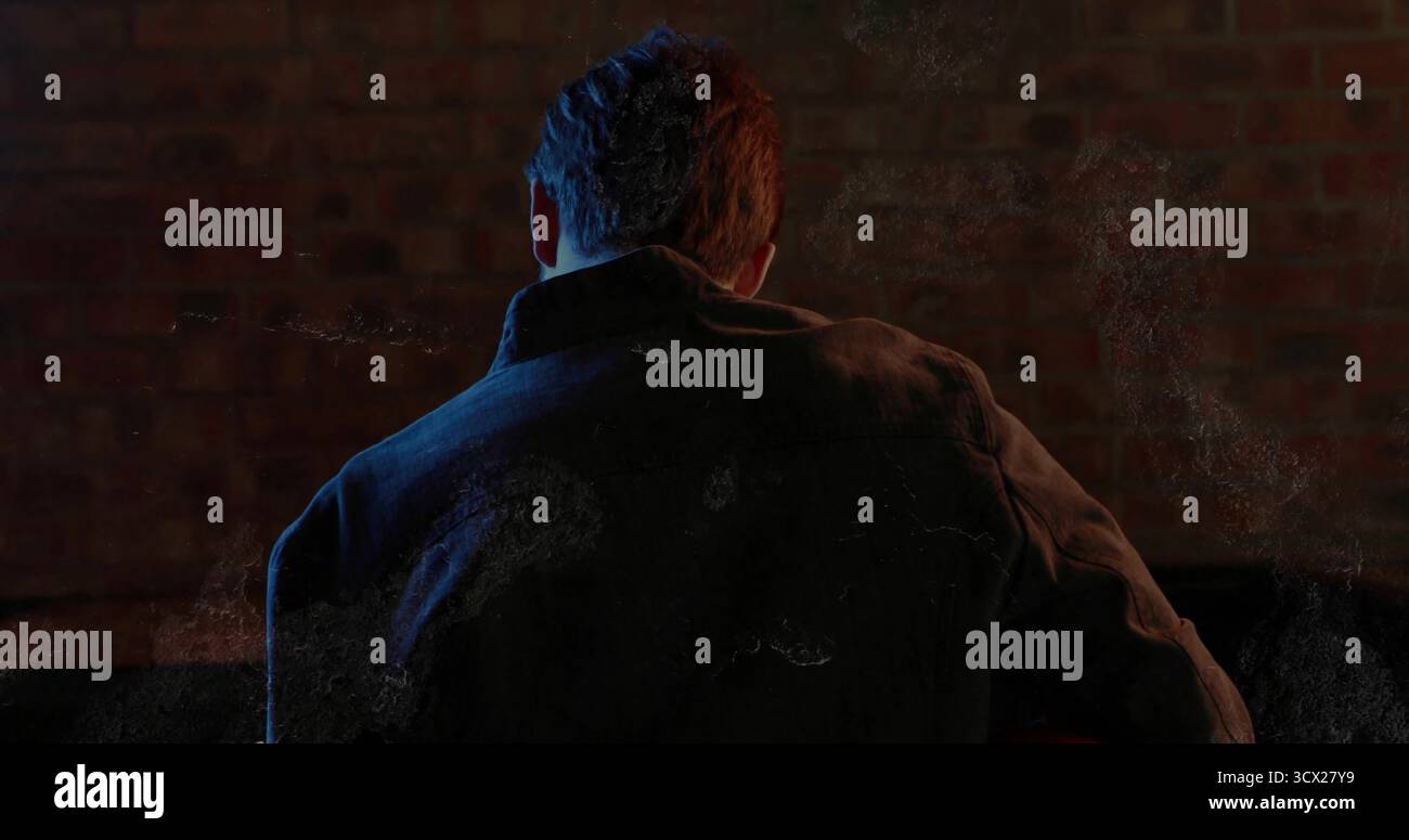 Uomo in piedi che indossa una giacca in tela scura contro le pareti in mattoni in studio, con illuminazione d'accento colorata Foto Stock