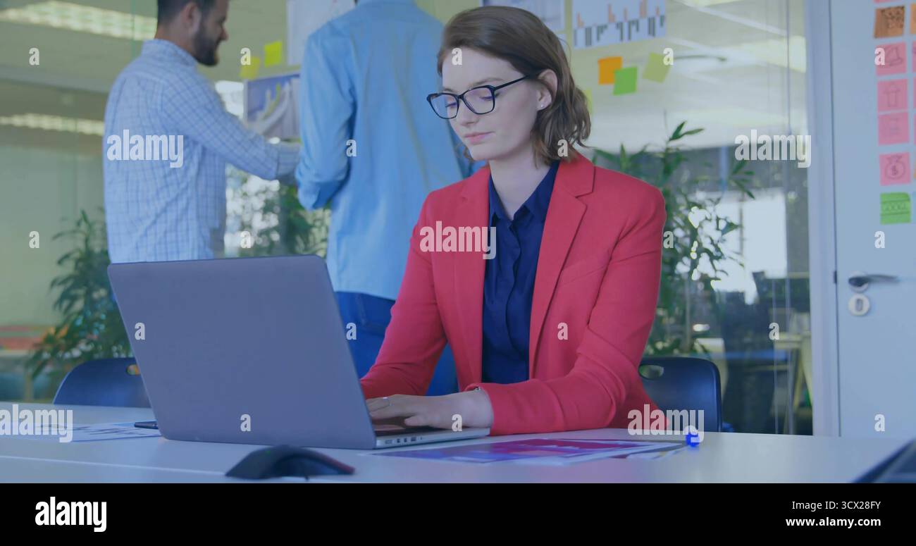 Donna che indossa un blazer rosso che scrive su un notebook in ufficio, appunti adesivi su una parete di vetro, documenti stampati Foto Stock