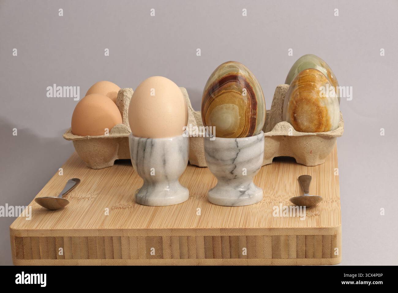 Natura morta con le uova. Tre uova reali e tre di pietra su un bordo di bambù con cucchiai inossidabili. Una delle uova si trova sempre nel porta uova. Foto Stock