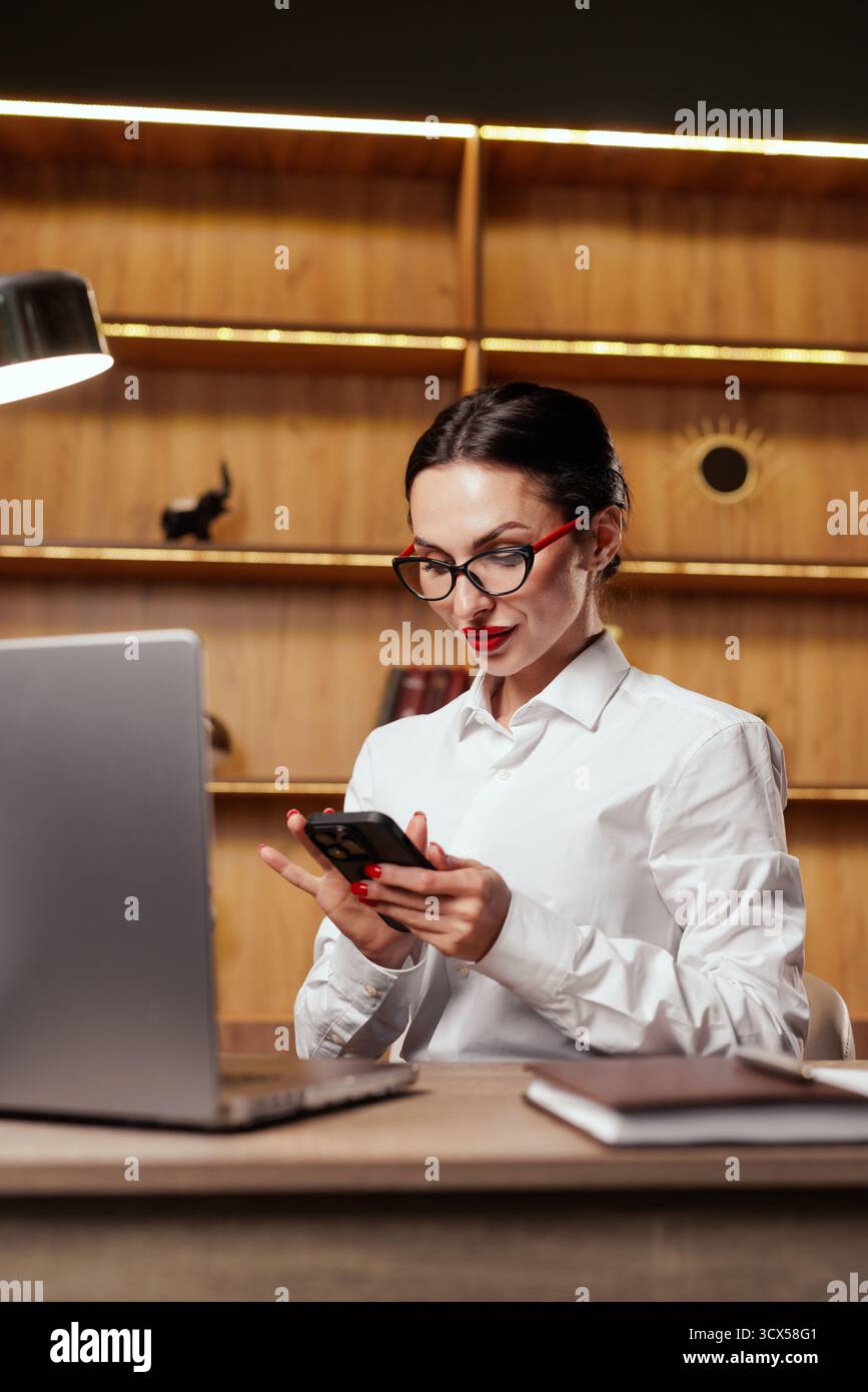 Una donna professionista controlla un telefono accanto al suo laptop, perfetto per concetti di messaggistica, funzionalità dell'app, verifica, contatto con i clienti e ufficio moderno Foto Stock