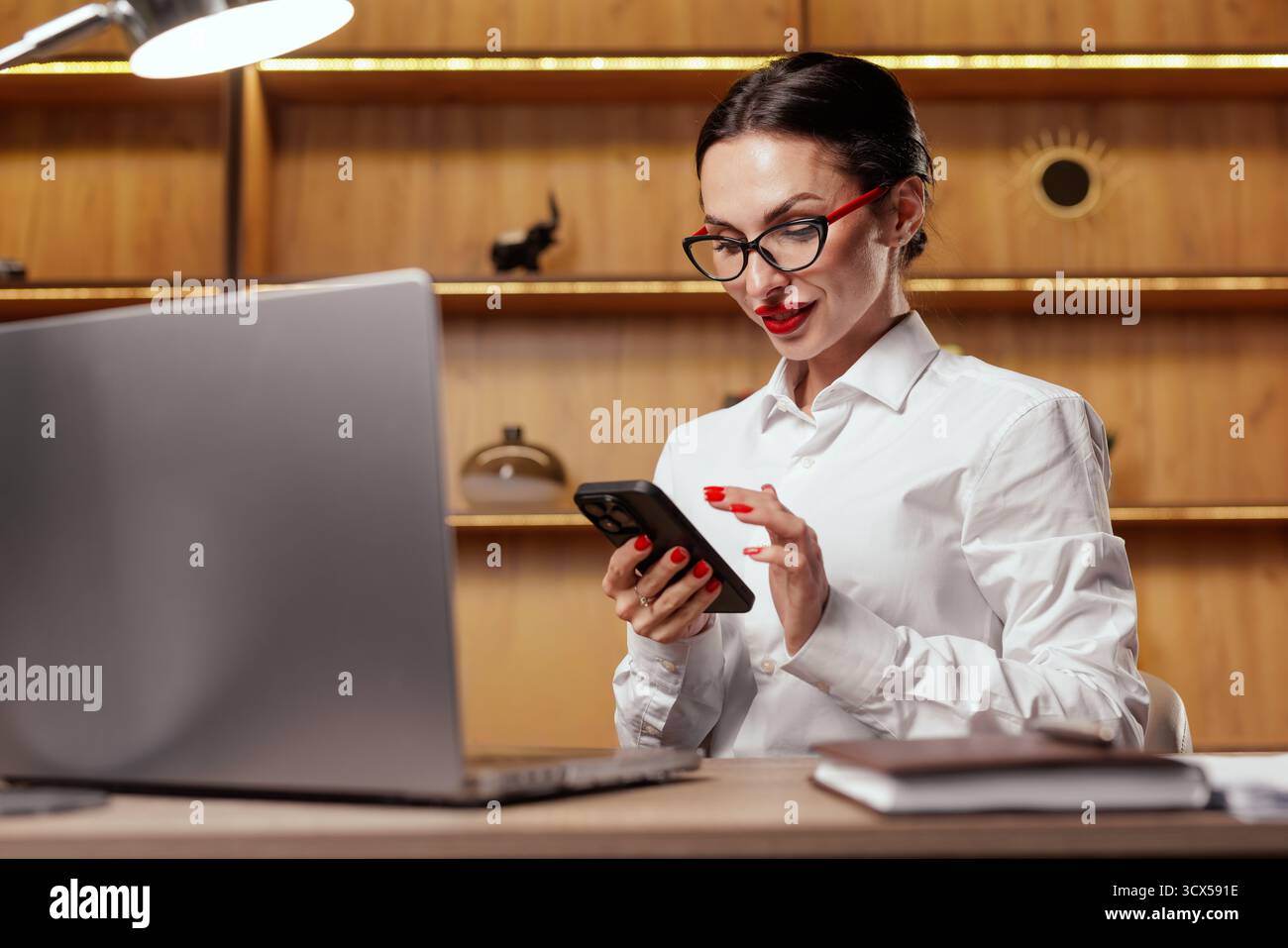 Una donna professionista controlla i messaggi al telefono mentre lavora su un laptop, ideale per applicazioni mobili, comunicazioni, login, verifica e flusso di lavoro dei clienti Foto Stock