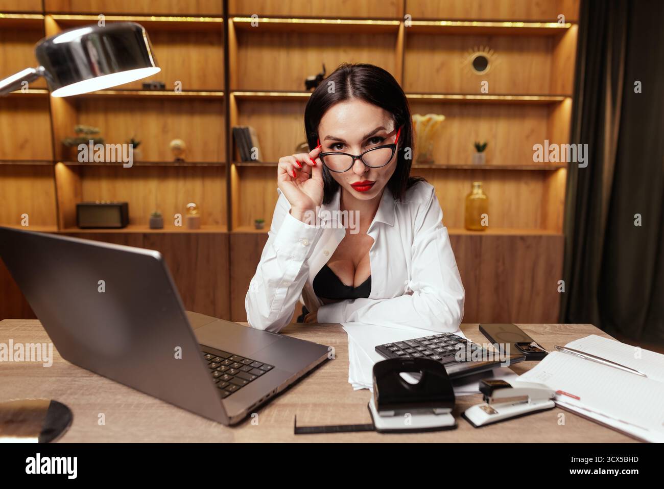 Una donna dirigente dà uno sguardo severo sugli occhiali alla scrivania dell'ufficio con laptop e documenti, simboleggiando autorità, disciplina e controllo Foto Stock