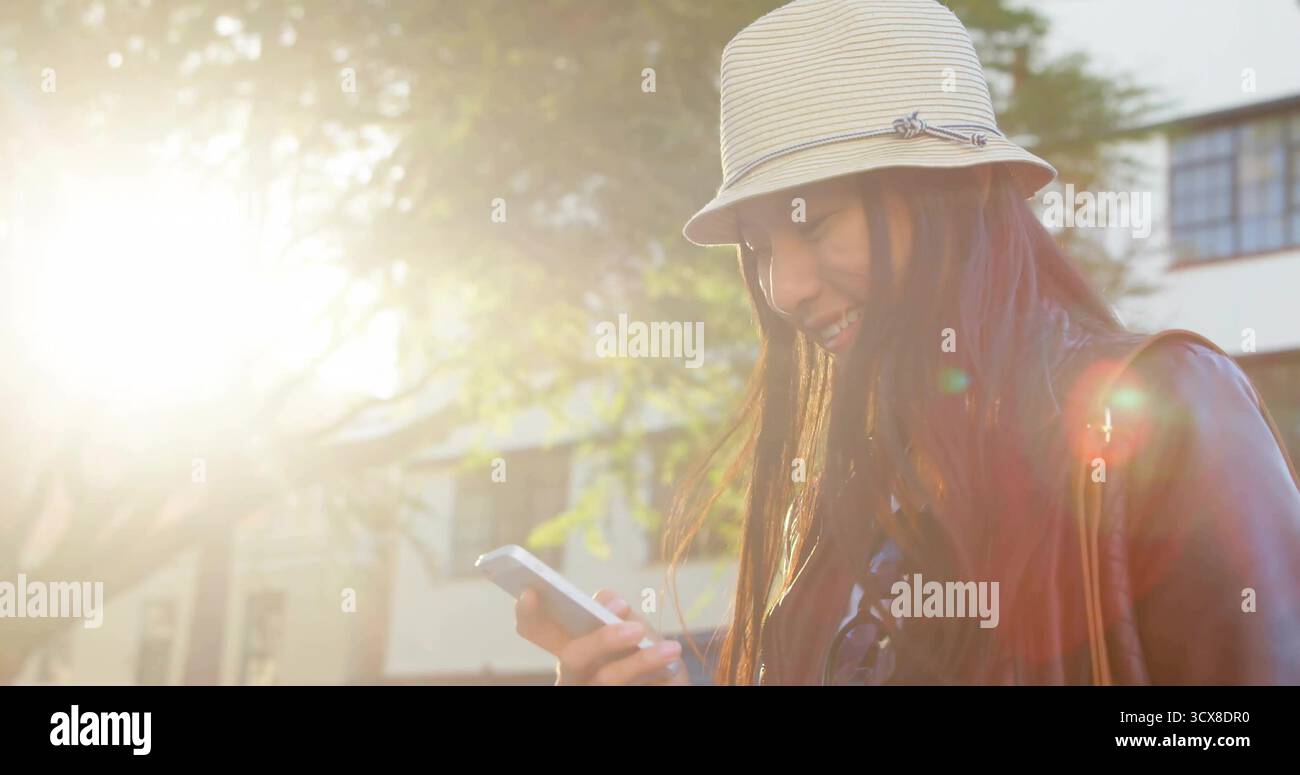 Donna sorridente che regge lo smartphone sul marciapiede illuminato dal sole, cappello a secchio a righe, giacca in pelle, spazio fotocopie Foto Stock