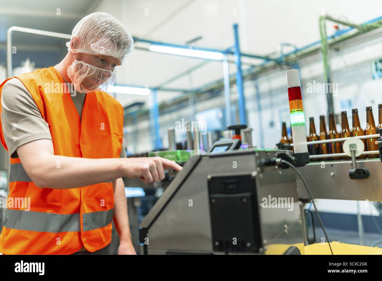 Lavoratore maschile in attrezzatura protettiva che supervisiona l'imbottigliamento e l'etichettatura automatizzati della birra su una linea di trasporto moderna e pulita, monitorando i macchinari per il controllo della qualità Foto Stock