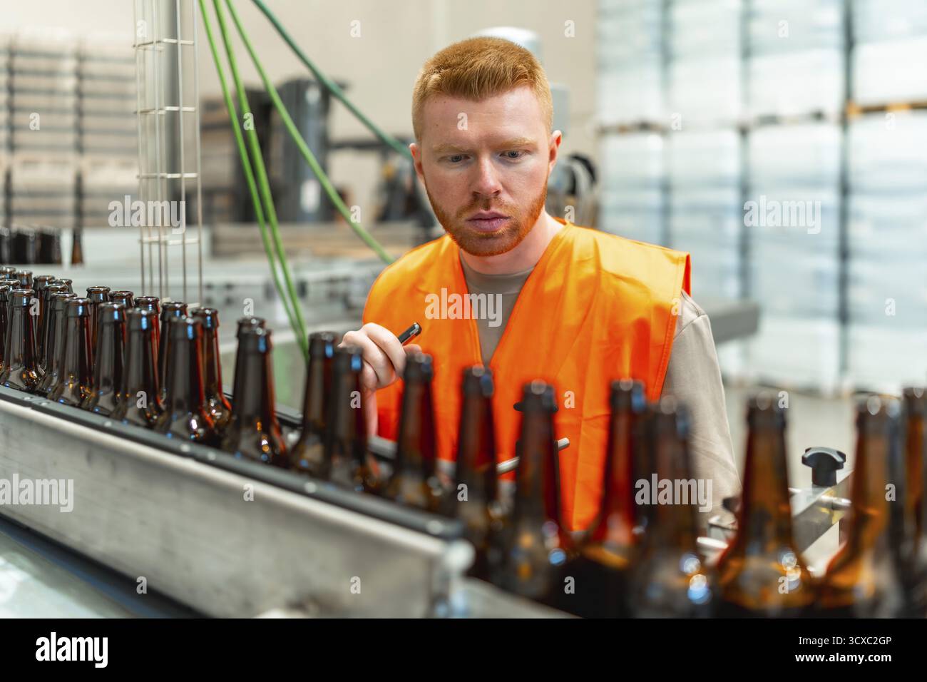 Uomo che indossa un giubbotto di sicurezza arancione che ispeziona le bottiglie di birra in vetro vuote che si spostano lungo un nastro trasportatore in una fabbrica di birra industriale, garantendo la qualità Foto Stock