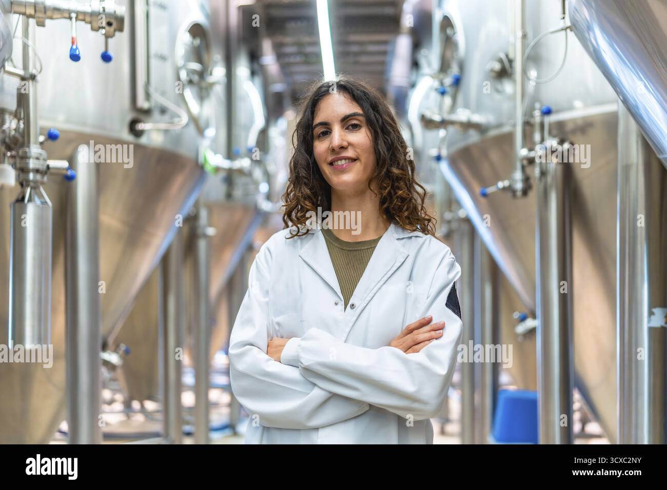 Donna che indossa un camice da laboratorio con le braccia incrociate in un birrificio industriale contemporaneo, circondato da un'ampia fermentazione in acciaio inox Foto Stock