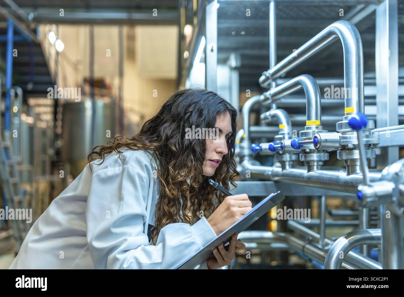 Donna che indossa un camice da laboratorio che controlla meticolosamente i tubi e le valvole industriali in una moderna fabbrica di bevande, eseguendo un controllo qualità dettagliato Foto Stock