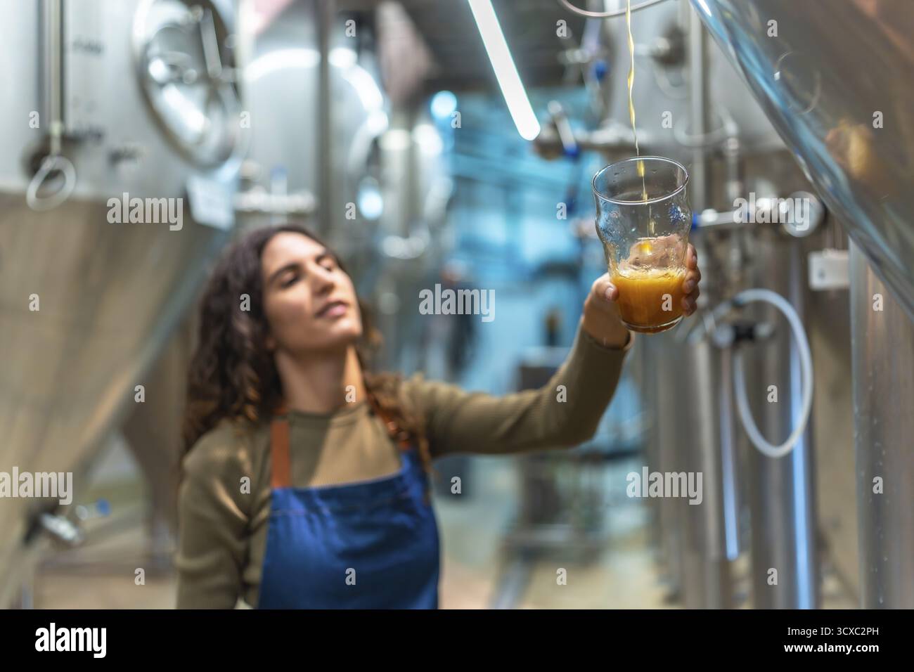 Una donna che indossa un grembiule, versa con cura birra artigianale appena prodotta in un bicchiere per un test di controllo qualità all'interno di un moderno birrificio con Foto Stock