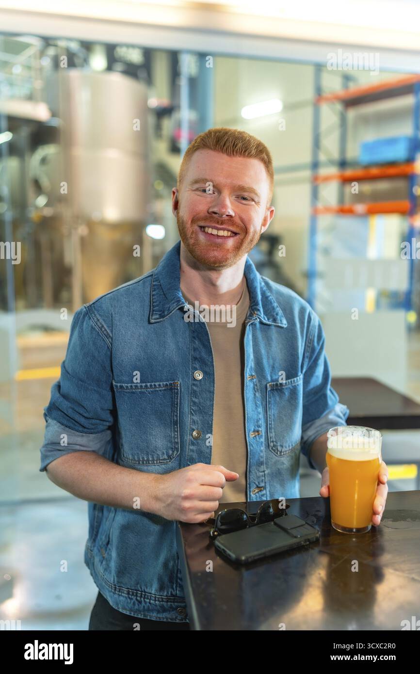 Uomo sorridente che si gusta un bicchiere di birra artigianale in una moderna birreria industriale. Indossa una giacca in denim e guarda la macchina fotografica con un Happy expres Foto Stock