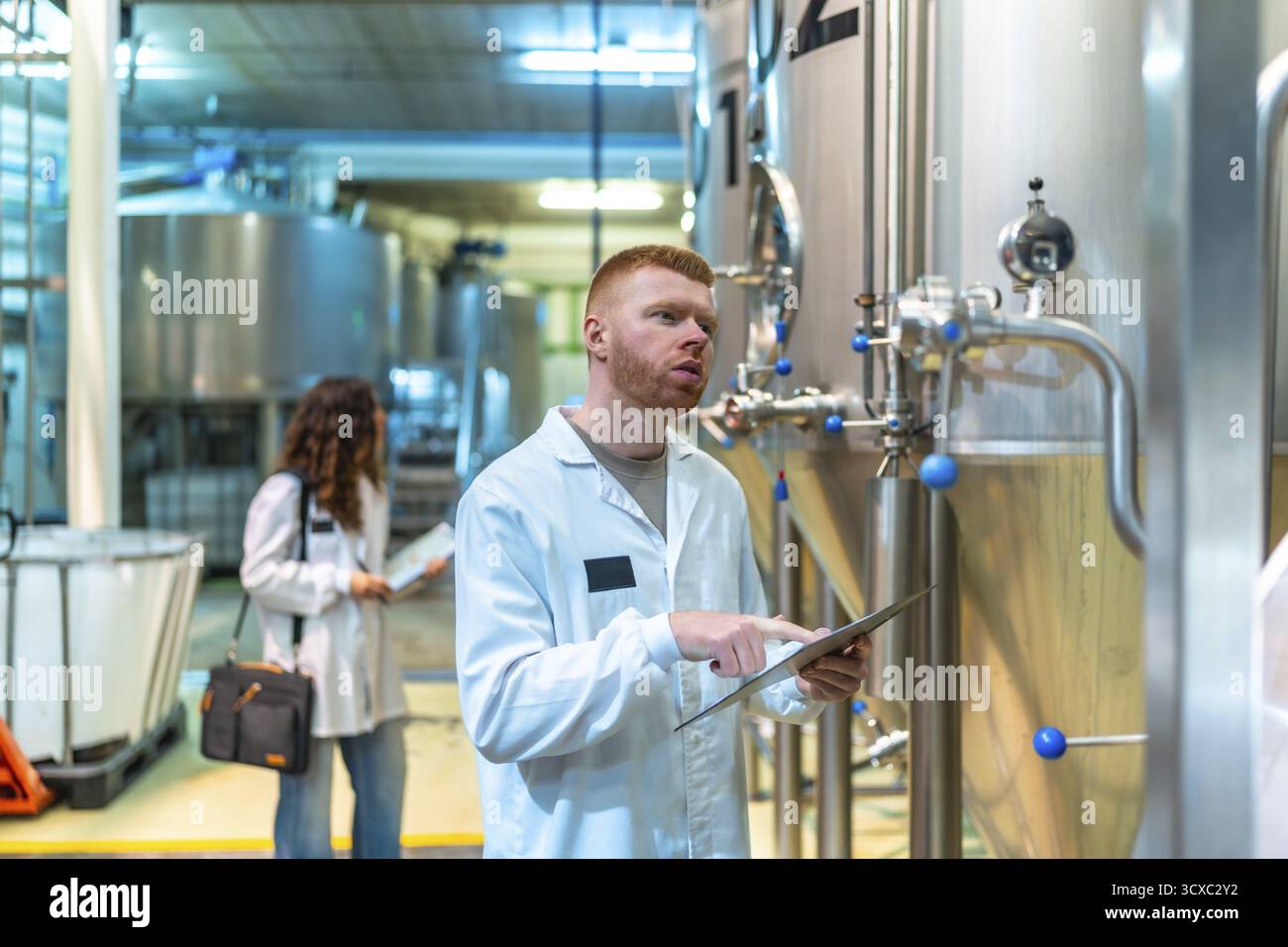 Il team di controllo qualità della birreria ispeziona i serbatoi di fermentazione industriale, un tecnico monitora i dati su un tablet mentre un altro lavoratore cammina tra le macchie Foto Stock
