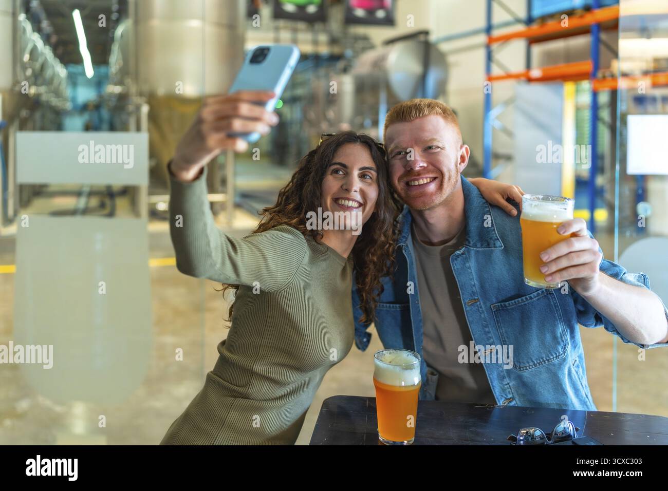 Giovane coppia sorridente per un selfie con birre artigianali in mano in una moderna sala di degustazione della birreria, godendosi un informale ritrovo nel fine settimana, degustando e condividendo Foto Stock