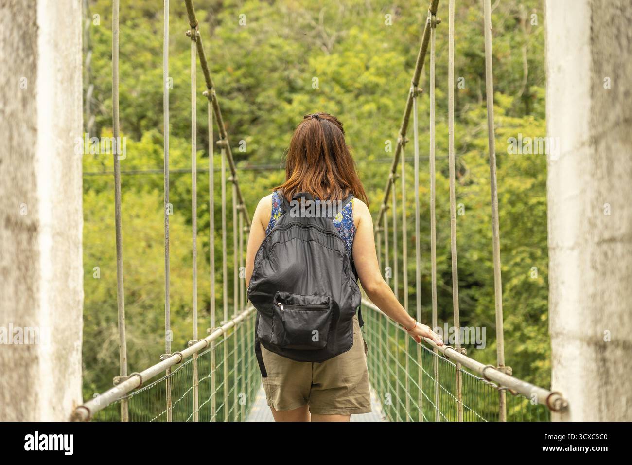 Turista donna con zaino che cammina su un ponte sospeso sul fiume sella, circondato da una lussureggiante foresta verde nelle asturie, in spagna, godendosi un'estate Foto Stock
