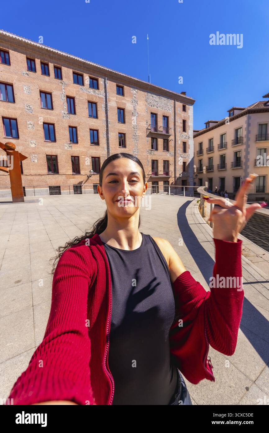 Giovane donna con i capelli scuri che posa per un selfie in una piazza della città, tenendo la mano in alto, con vecchi edifici europei e un cielo blu limpido nel backgrou Foto Stock