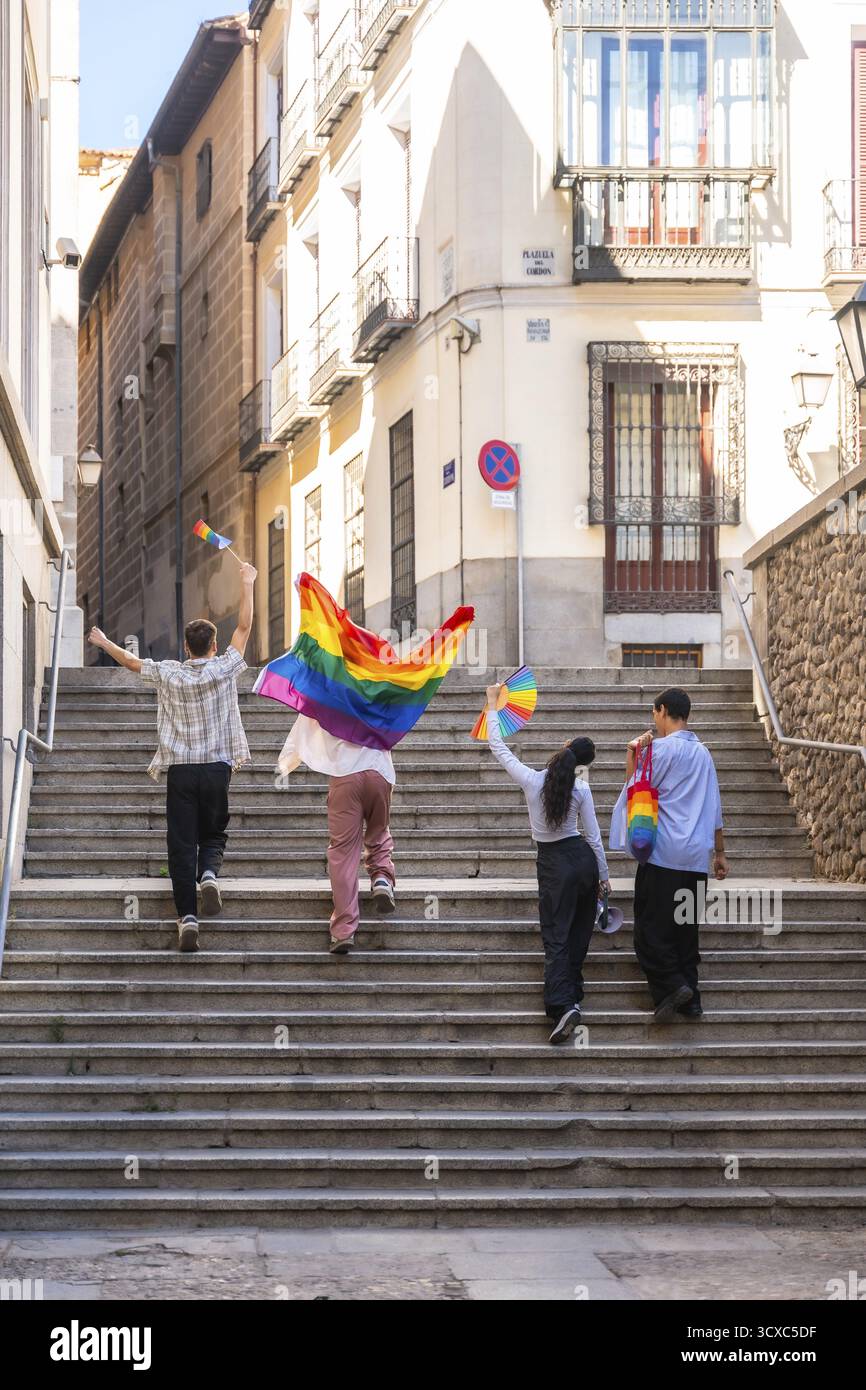 I giovani adulti camminano con gioia su scale di pietra in una città europea, portando bandiere arcobaleno e una borsa, celebrando la diversità e la lgbtq Plus commu Foto Stock