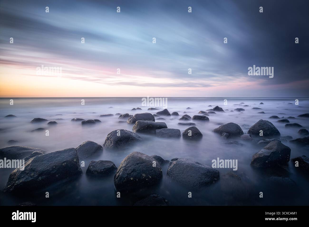 Costa rocciosa con massi lisci e arrotondati parzialmente sommersi da onde nebbiose al tramonto. Il cielo mostra striature di nuvole. Foto Stock