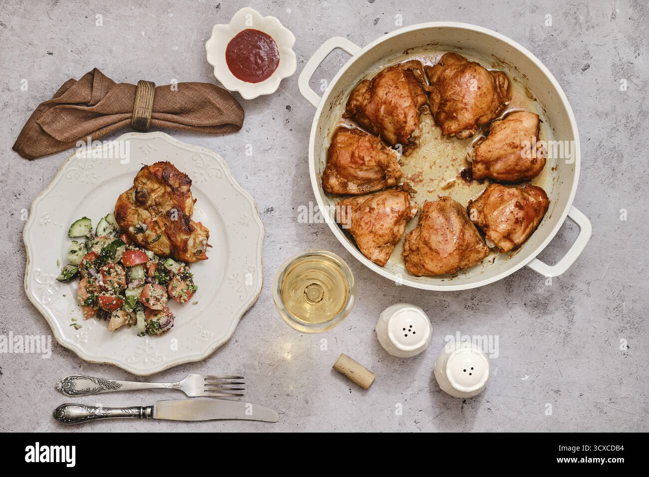 Un piatto ben cotto di cosce di pollo arrosto in una grande pentola, accompagnato da una vibrante insalata di cetrioli e pomodori su un piatto, accanto a un bicchiere di Foto Stock