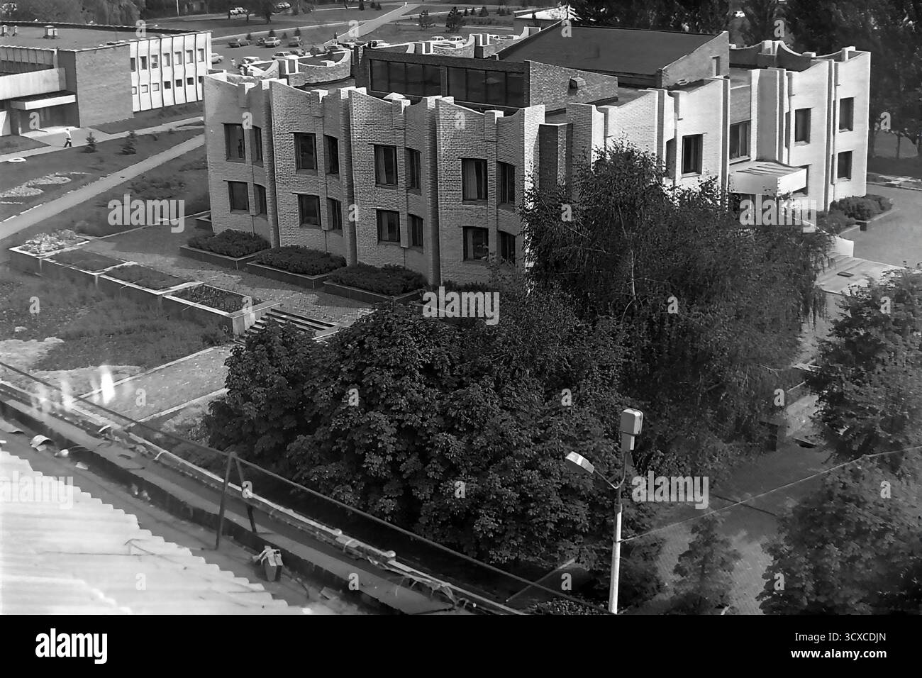 Una foto d'archivio ad alto angolo degli anni '1980 mostra la Biblioteca centrale della città in Piazza della Rivoluzione d'ottobre (ora Piazza Soborna) a Sloviansk. L'edificio e' un esempio impressionante di architettura modernista tardo-sovietica, notevole per la sua complessa facciata in mattoni e l'unica silhouette a castellata. Sotto, i pedoni passeggiano lungo un marciapiede circondato da alberi lussureggianti, che rappresentano un momento della vita quotidiana. Questa foto è una preziosa testimonianza dell'identità urbana e culturale di un tranquillo Donbas, che cattura l'atmosfera della città prima della guerra. Foto Stock