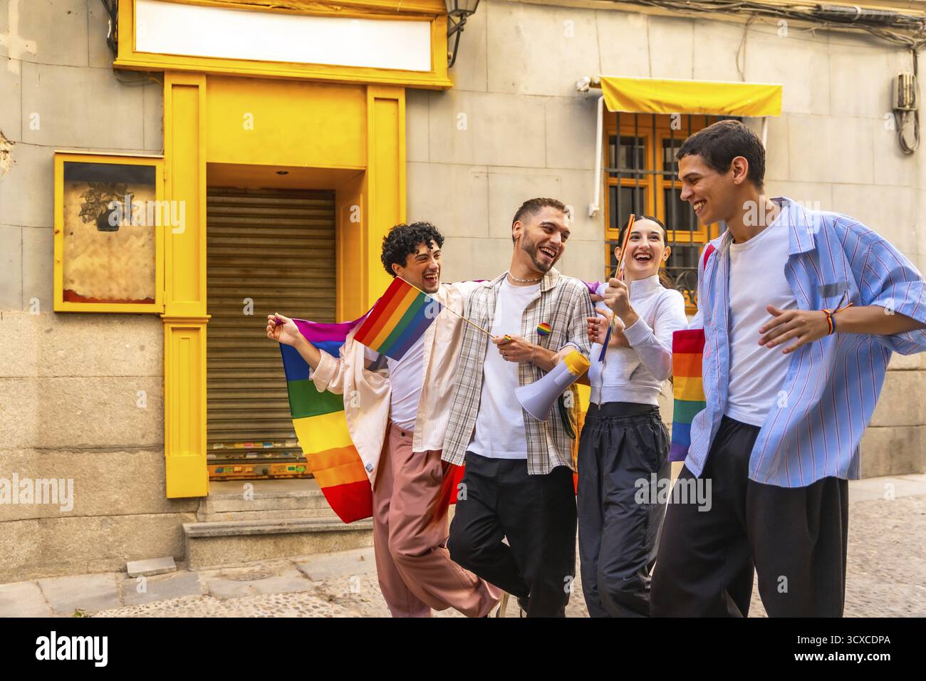 Giovani e variegati amici che celebrano la lgbtqia e l'orgoglio, camminano allegramente su una strada della città, portando bandiere arcobaleno e un megafono, abbracciando gioia e Foto Stock