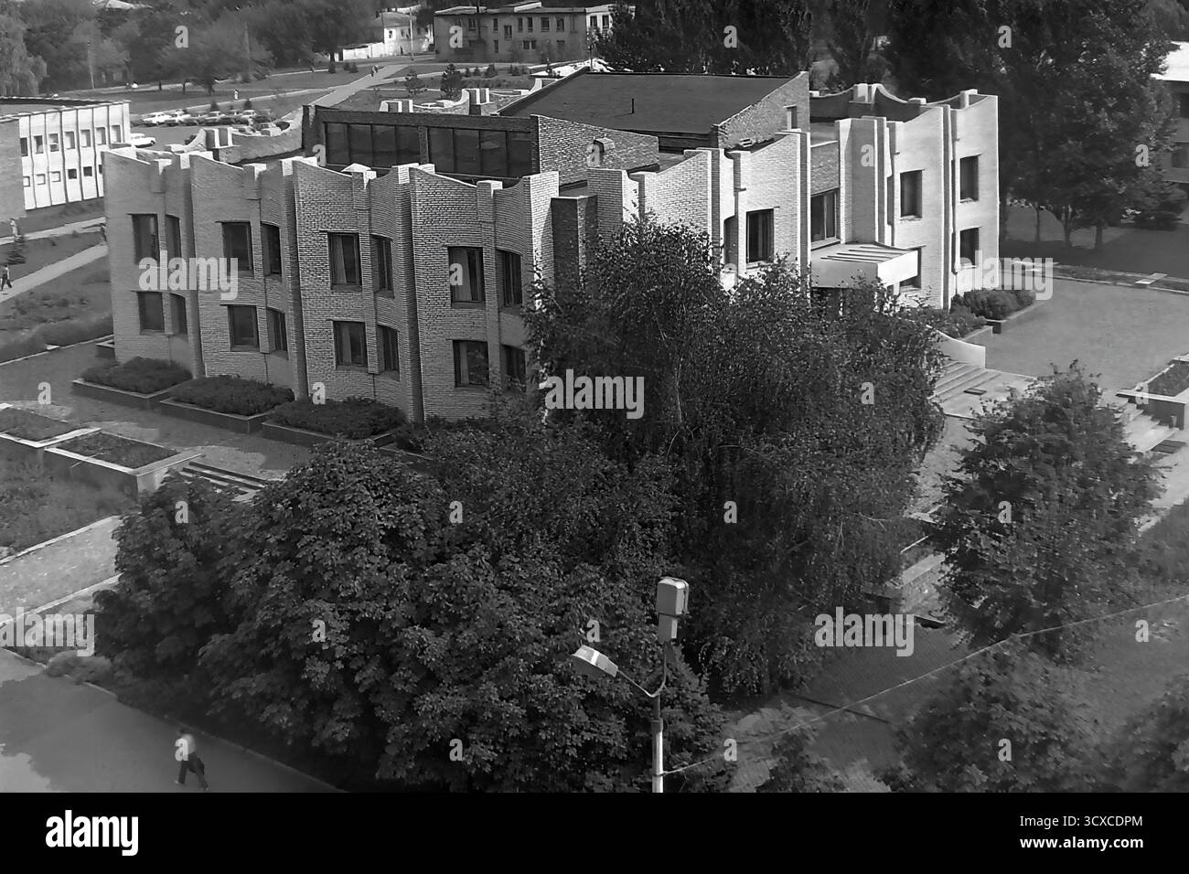 Una foto d'archivio ad alto angolo degli anni '1980 mostra la Biblioteca centrale della città in Piazza della Rivoluzione d'ottobre (ora Piazza Soborna) a Sloviansk. L'edificio e' un esempio impressionante di architettura modernista tardo-sovietica, notevole per la sua complessa facciata in mattoni e l'unica silhouette a castellata. Sotto, i pedoni passeggiano lungo un marciapiede circondato da alberi lussureggianti, che rappresentano un momento della vita quotidiana. Questa foto è una preziosa testimonianza dell'identità urbana e culturale di un tranquillo Donbas, che cattura l'atmosfera della città prima della guerra. Foto Stock