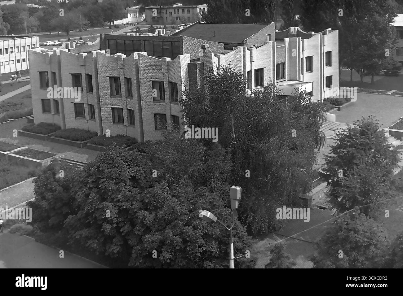 Una foto d'archivio ad alto angolo degli anni '1980 mostra la Biblioteca centrale della città in Piazza della Rivoluzione d'ottobre (ora Piazza Soborna) a Sloviansk. L'edificio e' un esempio impressionante di architettura modernista tardo-sovietica, notevole per la sua complessa facciata in mattoni e l'unica silhouette a castellata. Sotto, i pedoni passeggiano lungo un marciapiede circondato da alberi lussureggianti, che rappresentano un momento della vita quotidiana. Questa foto è una preziosa testimonianza dell'identità urbana e culturale di un tranquillo Donbas, che cattura l'atmosfera della città prima della guerra. Foto Stock