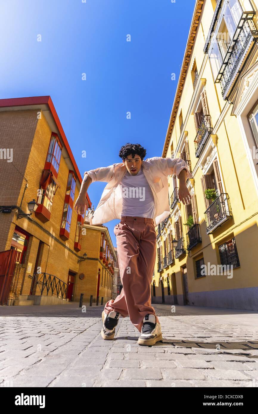 Giovane uomo elegante con un'espressione intensa che esegue una dinamica danza di strada su ciottoli in un vivace quartiere cittadino europeo, a tutta lunghezza, sunn Foto Stock