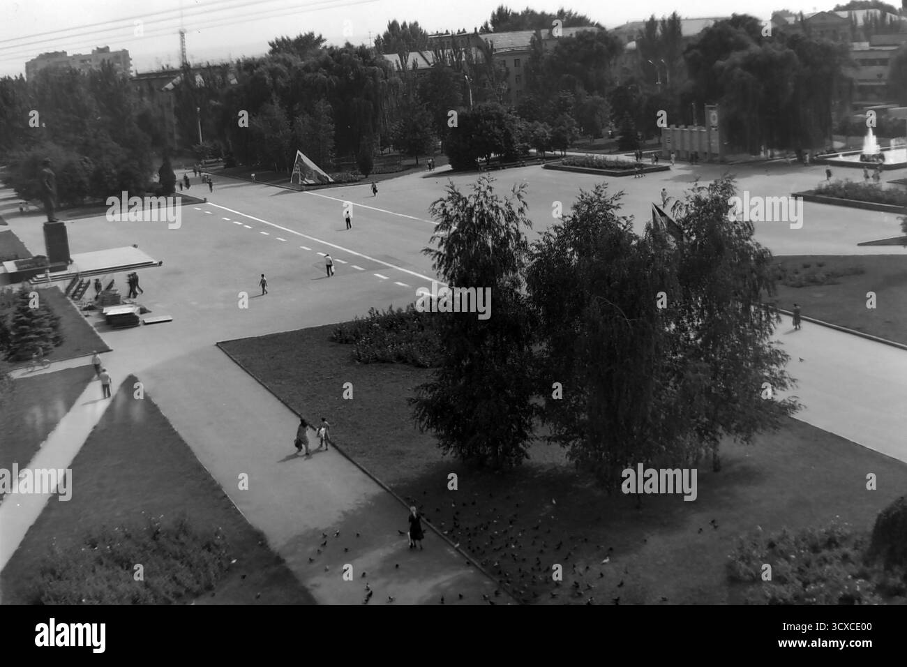 Una foto d'archivio ad alto angolo degli anni '1980 mostra l'ampia Piazza della Rivoluzione d'ottobre (ora Piazza Soborna) a Sloviansk, RSS Ucraina. Questo spazio pubblico centrale presenta un importante monumento sulla sinistra, fontane e bandiere sovietiche. I pedoni passeggiano tranquillamente attraverso l'area aperta, mentre uno stormo di piccioni si riunisce in primo piano, creando un'atmosfera vivace. Questa immagine cattura una scena tipica della vita quotidiana e del tempo libero in una città sovietica, un prezioso resoconto storico di un tranquillo Donbas prima della guerra Foto Stock