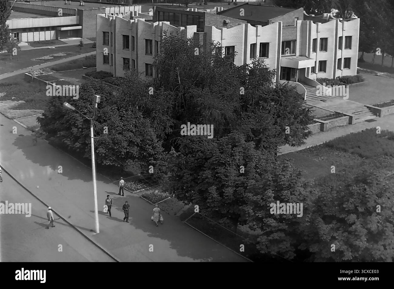 Una foto d'archivio ad alto angolo degli anni '1980 mostra la Biblioteca centrale della città in Piazza della Rivoluzione d'ottobre (ora Piazza Soborna) a Sloviansk. L'edificio e' un esempio impressionante di architettura modernista tardo-sovietica, notevole per la sua complessa facciata in mattoni e l'unica silhouette a castellata. Sotto, i pedoni passeggiano lungo un marciapiede circondato da alberi lussureggianti, che rappresentano un momento della vita quotidiana. Questa foto è una preziosa testimonianza dell'identità urbana e culturale di un tranquillo Donbas, che cattura l'atmosfera della città prima della guerra. Foto Stock