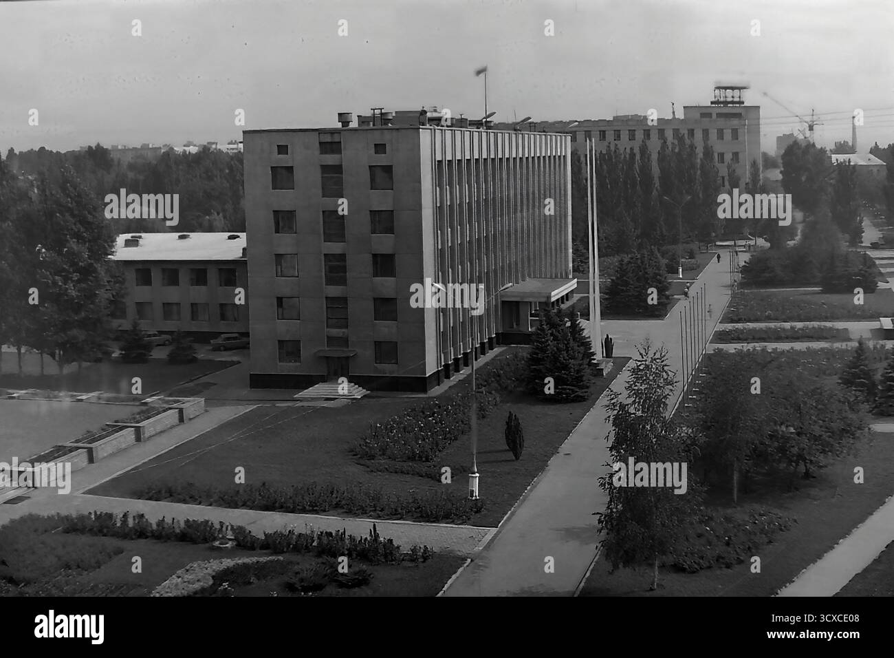 Una foto d'archivio ad alto angolo degli anni '1980 cattura il Comitato Esecutivo della città di Sloviansk in Piazza della Rivoluzione d'ottobre (ora Piazza Soborna). Il design rigoroso e funzionalista dell'edificio è un ottimo esempio di architettura modernista sovietica. La piazza circostante è meticolosamente paesaggizzata con passaggi geometrici, aiuole e prati curati, dove passeggiano alcuni pedoni. Questa immagine conserva un momento di ordinata vita pubblica in un tranquillo Donbas, riflettendo l'ambiente urbano strutturato dell'epoca prima della guerra Foto Stock