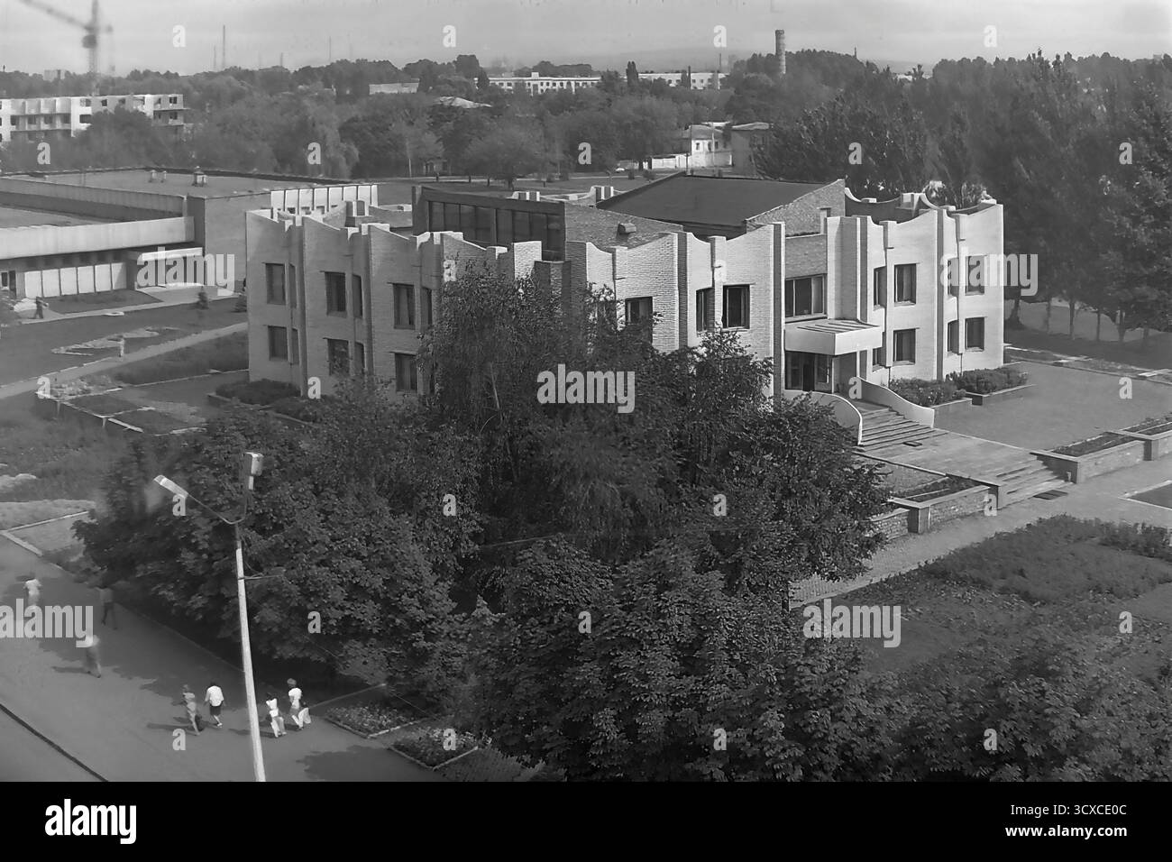 Una foto d'archivio ad alto angolo degli anni '1980 mostra la Biblioteca centrale della città in Piazza della Rivoluzione d'ottobre (ora Piazza Soborna) a Sloviansk. L'edificio e' un esempio impressionante di architettura modernista tardo-sovietica, notevole per la sua complessa facciata in mattoni e l'unica silhouette a castellata. Sotto, i pedoni passeggiano lungo un marciapiede circondato da alberi lussureggianti, che rappresentano un momento della vita quotidiana. Questa foto è una preziosa testimonianza dell'identità urbana e culturale di un tranquillo Donbas, che cattura l'atmosfera della città prima della guerra. Foto Stock