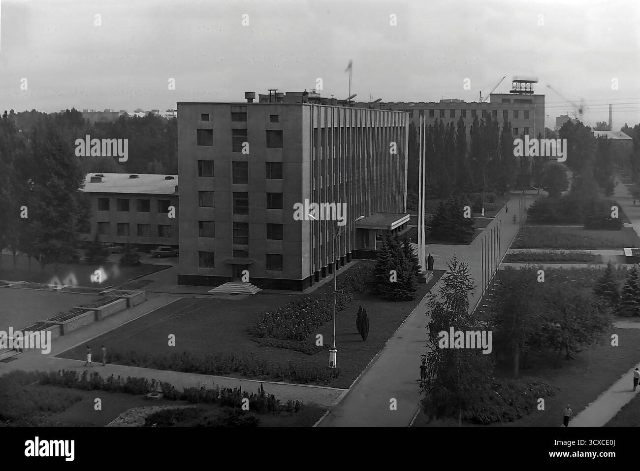 Una foto d'archivio ad alto angolo degli anni '1980 cattura il Comitato Esecutivo della città di Sloviansk in Piazza della Rivoluzione d'ottobre (ora Piazza Soborna). Il design rigoroso e funzionalista dell'edificio è un ottimo esempio di architettura modernista sovietica. La piazza circostante è meticolosamente paesaggizzata con passaggi geometrici, aiuole e prati curati, dove passeggiano alcuni pedoni. Questa immagine conserva un momento di ordinata vita pubblica in un tranquillo Donbas, riflettendo l'ambiente urbano strutturato dell'epoca prima della guerra Foto Stock