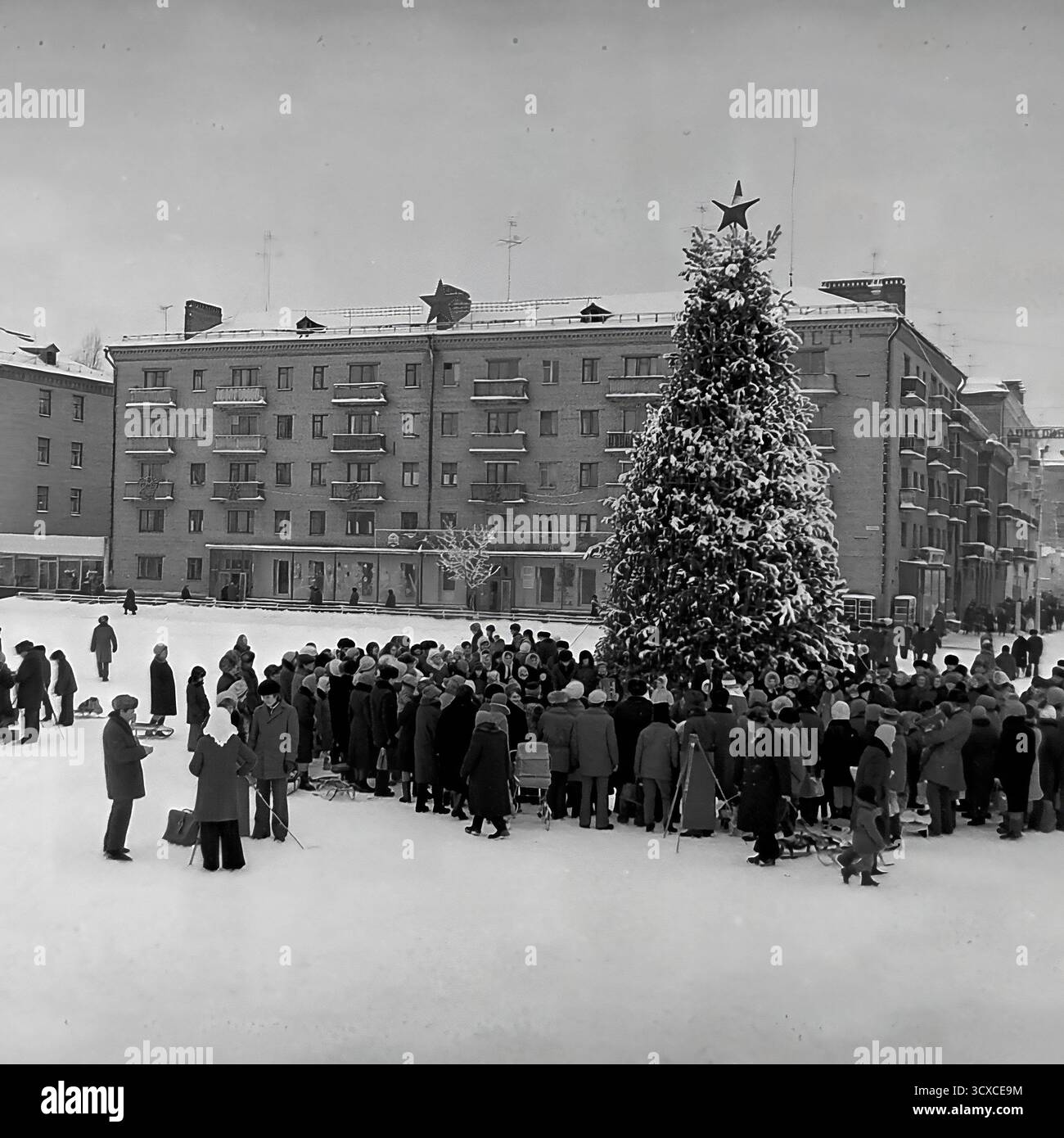 Un'ampia vista d'archivio dall'inverno 1976 mostra una celebrazione pubblica in Piazza della Rivoluzione d'ottobre (ora Piazza Soborna) a Sloviansk. Una grande folla di adulti e bambini si raduna intorno a un festoso albero di Capodanno sormontato da una stella sovietica. Uno striscione per il prossimo XXV Congresso del CPSU si estende dall'altra parte della strada sullo sfondo, un segno storico chiave dell'epoca. Questa foto cattura vividamente un momento di vita comunitaria e festività ufficiale sovietica, uno sguardo toccante allo spirito pubblico di un pacifico Donba prima della guerra Foto Stock