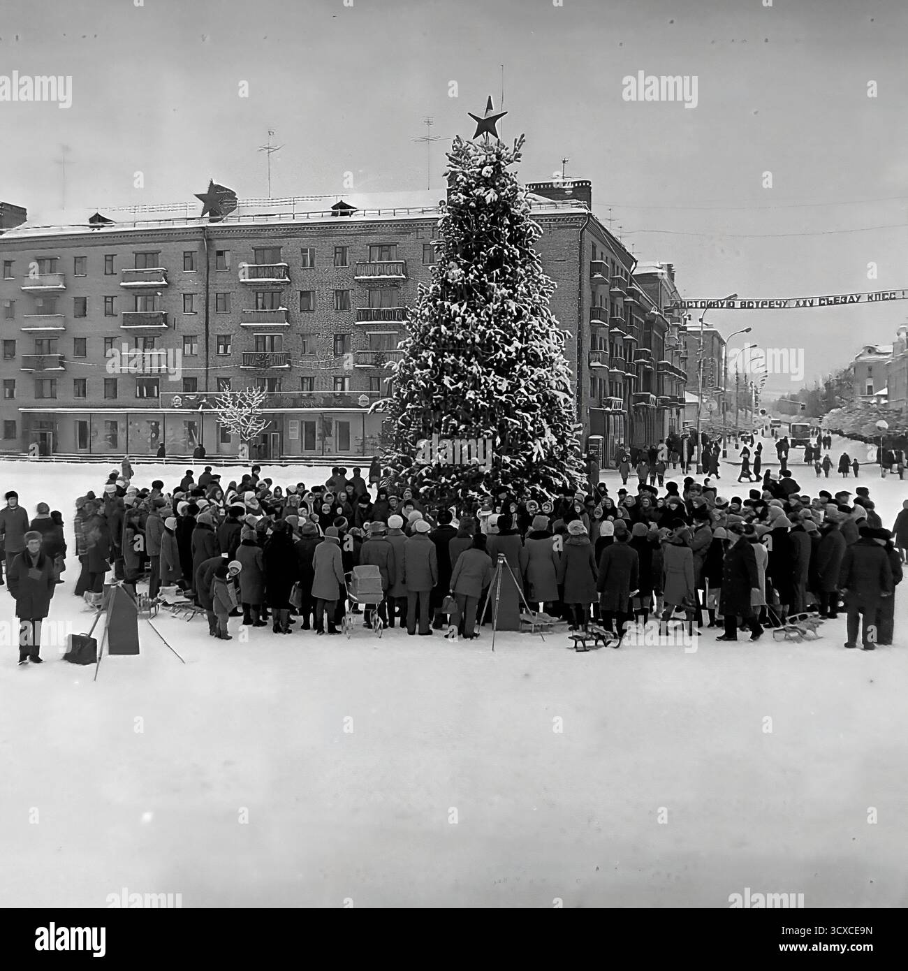 Un'ampia vista d'archivio dall'inverno 1976 mostra una celebrazione pubblica in Piazza della Rivoluzione d'ottobre (ora Piazza Soborna) a Sloviansk. Una grande folla di adulti e bambini si raduna intorno a un festoso albero di Capodanno sormontato da una stella sovietica. Uno striscione per il prossimo XXV Congresso del CPSU si estende dall'altra parte della strada sullo sfondo, un segno storico chiave dell'epoca. Questa foto cattura vividamente un momento di vita comunitaria e festività ufficiale sovietica, uno sguardo toccante allo spirito pubblico di un pacifico Donba prima della guerra Foto Stock