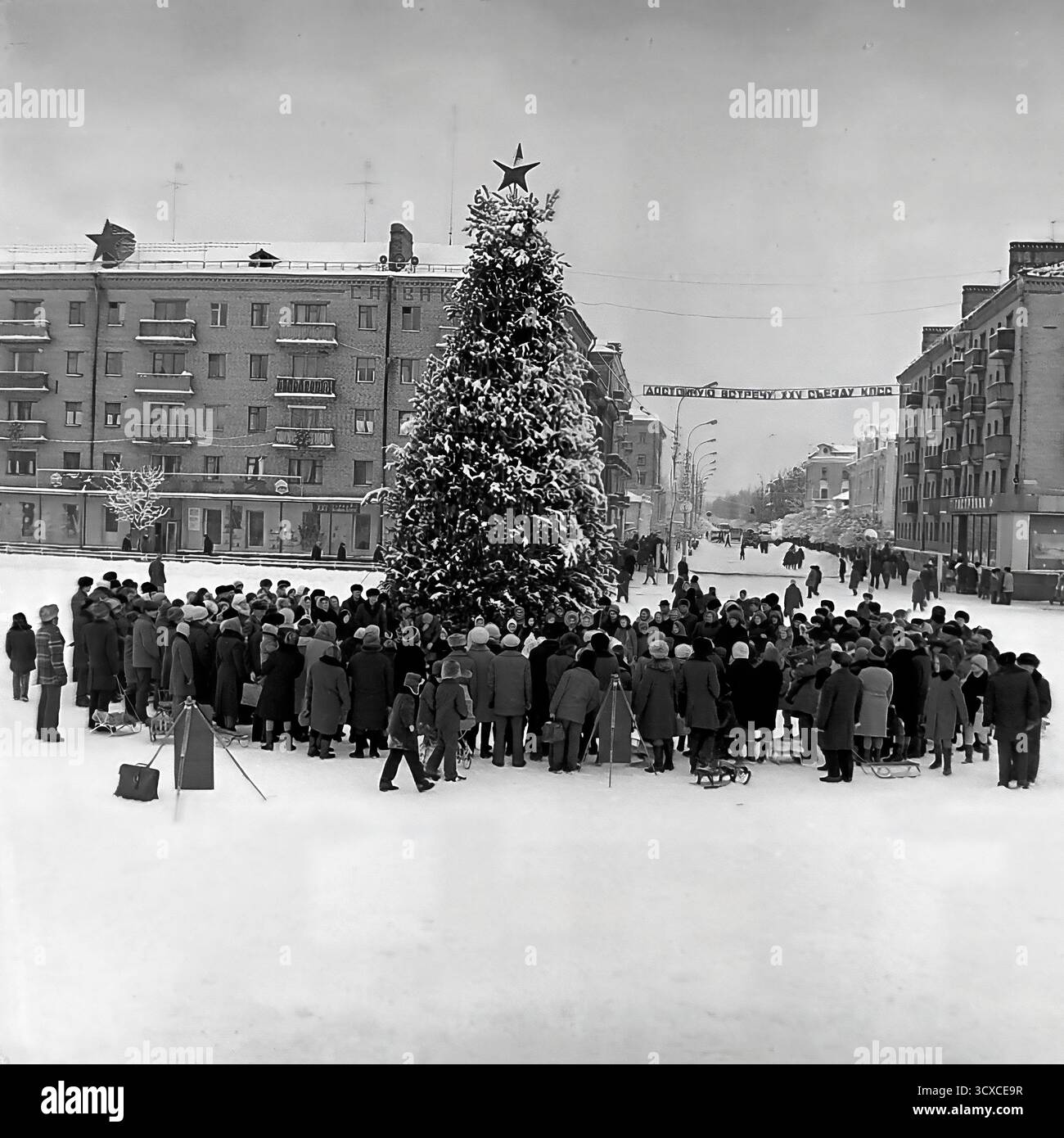 Un'ampia vista d'archivio dall'inverno 1976 mostra una celebrazione pubblica in Piazza della Rivoluzione d'ottobre (ora Piazza Soborna) a Sloviansk. Una grande folla di adulti e bambini si raduna intorno a un festoso albero di Capodanno sormontato da una stella sovietica. Uno striscione per il prossimo XXV Congresso del CPSU si estende dall'altra parte della strada sullo sfondo, un segno storico chiave dell'epoca. Questa foto cattura vividamente un momento di vita comunitaria e festività ufficiale sovietica, uno sguardo toccante allo spirito pubblico di un pacifico Donba prima della guerra Foto Stock
