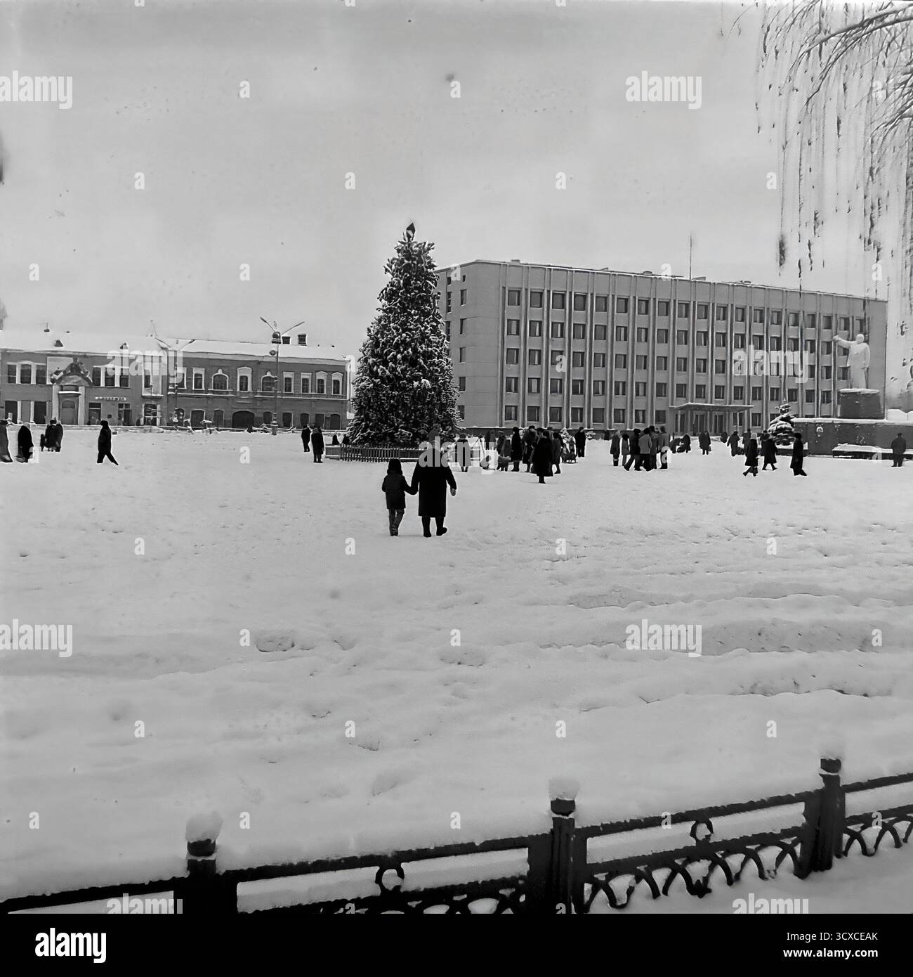 Una suggestiva foto d'archivio dell'inverno del 1976 mostra una piazza della Rivoluzione d'ottobre ricoperta di neve (ora Piazza Soborna) a Sloviansk. Un genitore e un bambino camminano mano nella mano verso l'albero centrale di Capodanno. Sullo sfondo si trovano l'imponente edificio del Comitato Esecutivo cittadino e il monumento Lenin, che definisce il paesaggio dell'era sovietica. La scena cattura un momento tranquillo della vita familiare in un grande spazio pubblico, un commovente record di routine quotidiana in un pacifico Donba prima della guerra Foto Stock