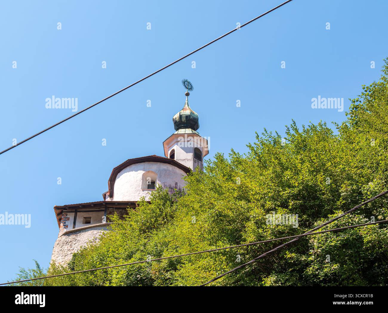 Kamnik, Slovenia - 10 agosto 2025: Una chiesa storica con una cupola verde e una croce, parzialmente oscurata dagli alberi, sotto un cielo azzurro. Linee elettriche cro Foto Stock Kamnik, Slovenia - 10 agosto 2025: Una chiesa storica con una cupola verde e una croce, parzialmente oscurata dagli alberi, sotto un cielo azzurro. Linee elettriche cro Foto Stock
