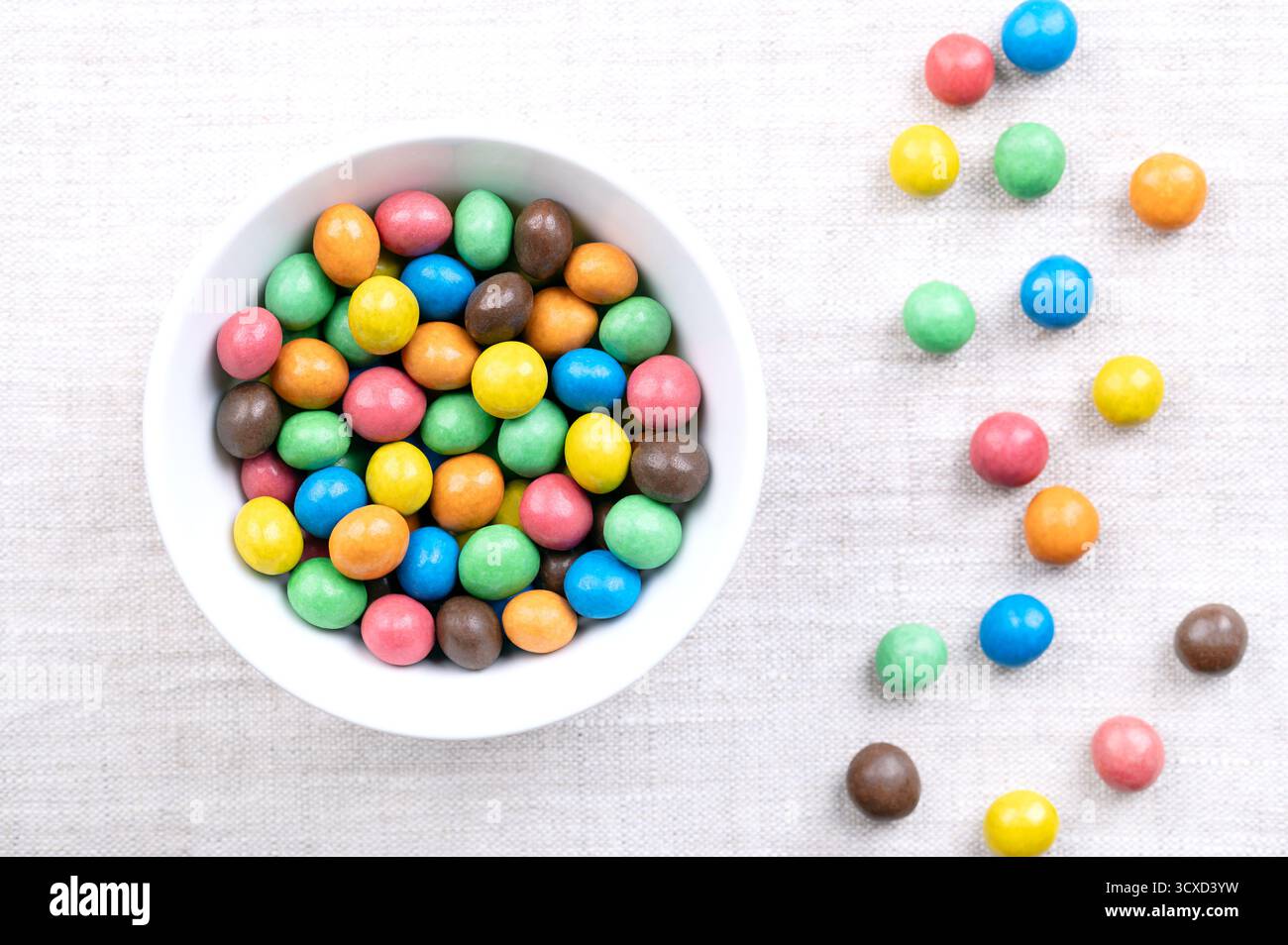 Colorate dragee al cioccolato ricoperte di zucchero, in una ciotola bianca, su lino. Dolciumi al cioccolato Dragee, costituiti da un guscio di caramelle multicolore. Foto Stock