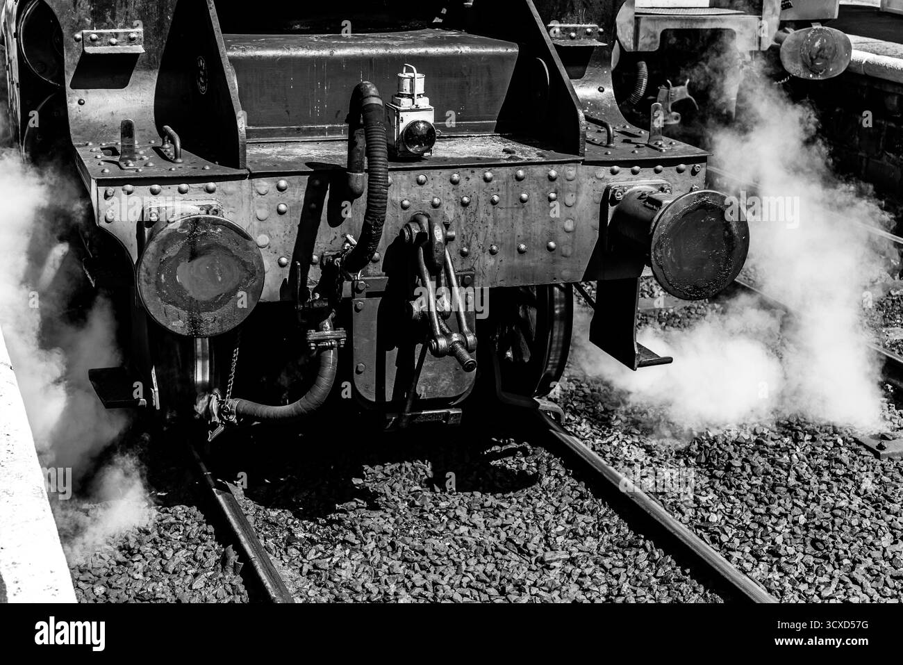 Primo piano bianco e nero di un buffer di locomotiva a vapore e giunti che rilasciano vapore in una stazione ferroviaria d'epoca Foto Stock
