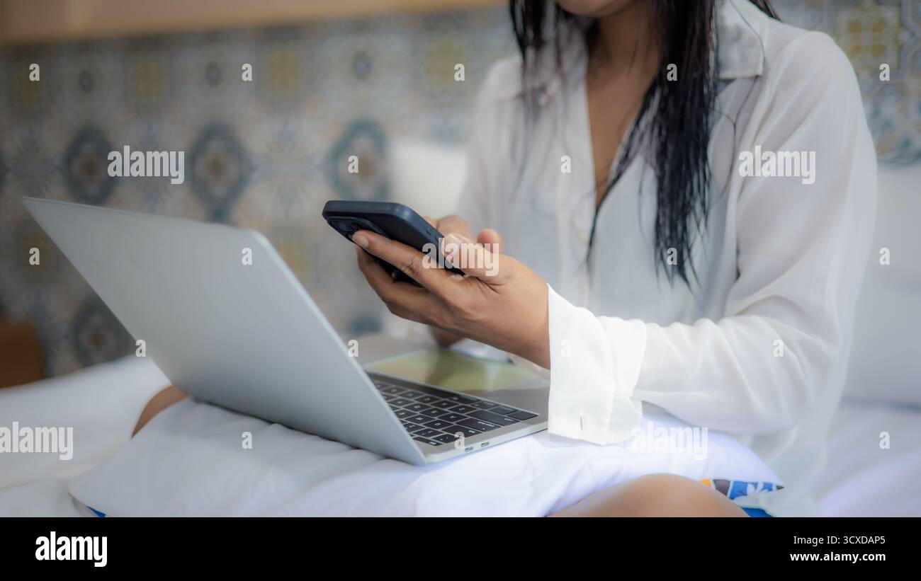 Primo piano di una donna che utilizza smartphone e computer portatile mentre lavora a distanza a letto. Concetto moderno di stile di vita nomade digitale, freelance o lavoro da qualsiasi luogo. Foto Stock