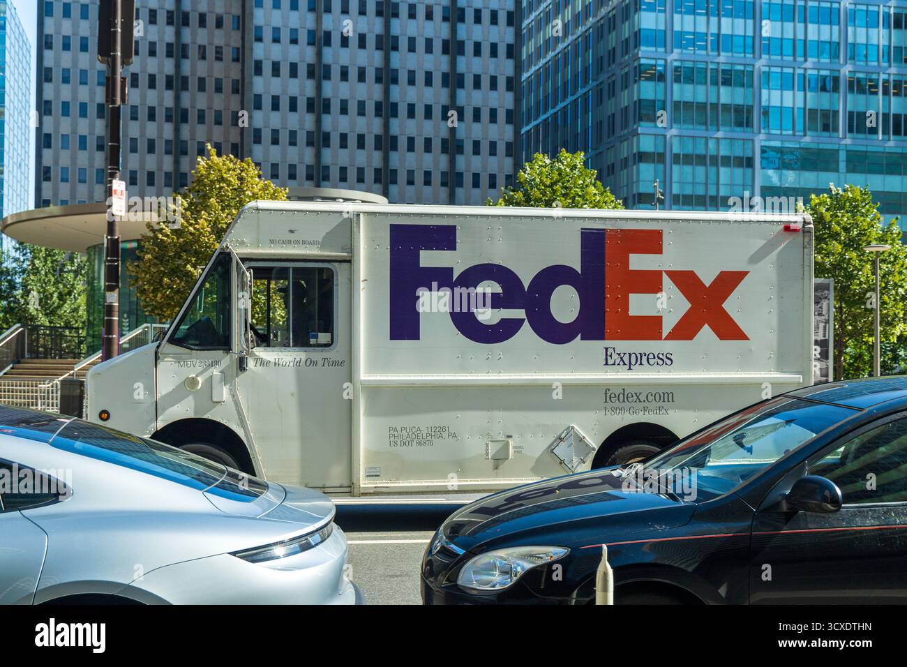 Un camion Fedex sta guidando lungo una strada trafficata. Il veicolo è bianco e presenta un logo viola e arancione sul lato. La strada è piena di auto, incluso Foto Stock Un camion Fedex sta guidando lungo una strada trafficata. Il veicolo è bianco e presenta un logo viola e arancione sul lato. La strada è piena di auto, incluso Foto Stock