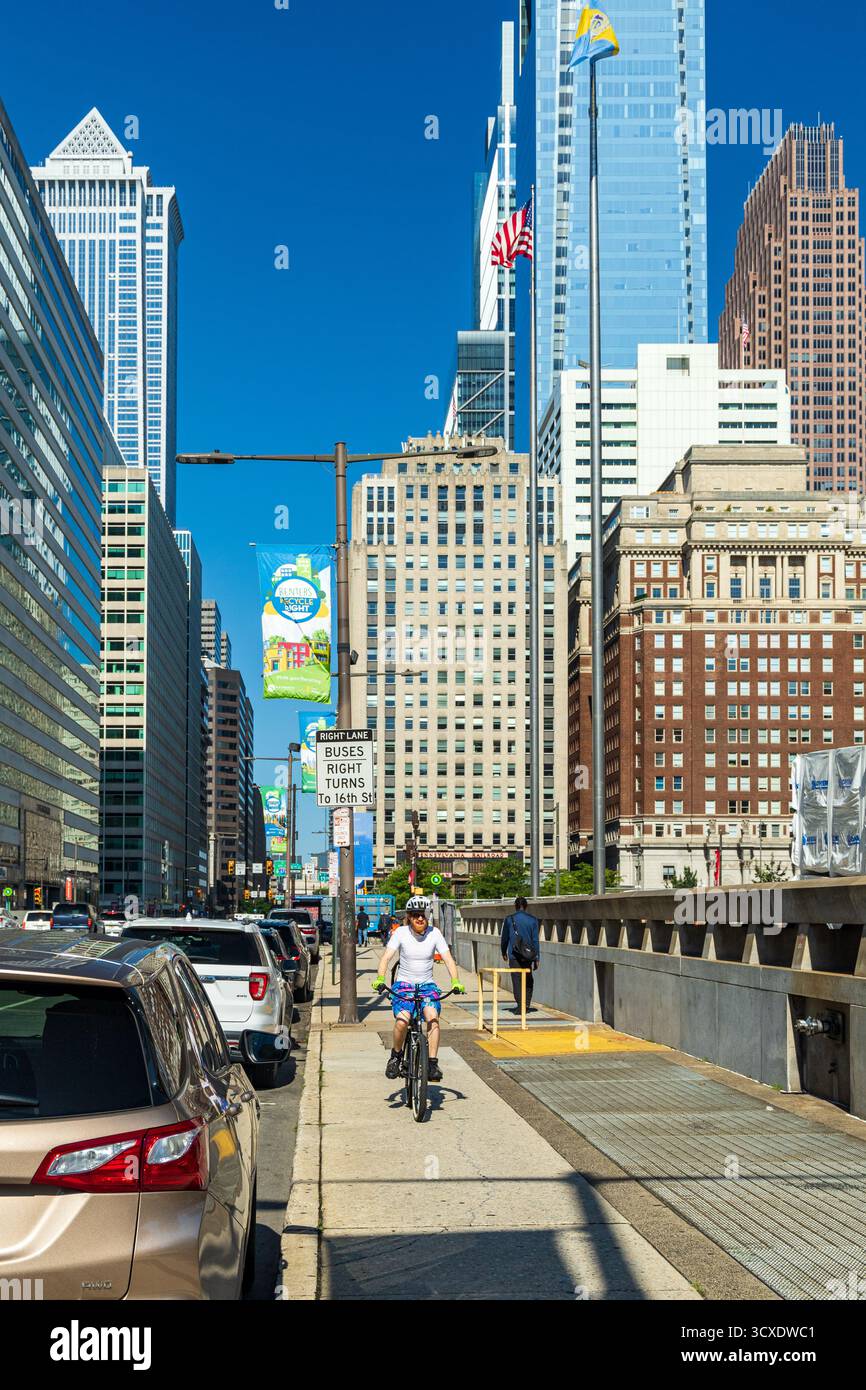 Un uomo corre in bicicletta lungo una strada cittadina. La strada è piena di auto e camion e ci sono diversi pedoni che camminano lungo il marciapiede. La S Foto Stock Un uomo corre in bicicletta lungo una strada cittadina. La strada è piena di auto e camion e ci sono diversi pedoni che camminano lungo il marciapiede. La S Foto Stock