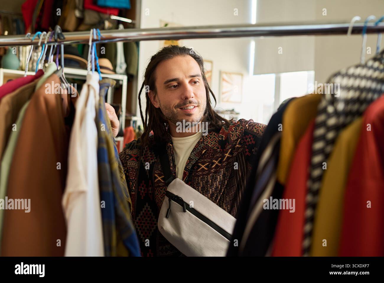 Sorridente giovane uomo caucasico che sfoglia il portapacchi, seleziona capi nel negozio di articoli sportivi, espressione mirata, capelli lunghi, in piedi tra abiti assortiti, shopping di moda di seconda mano Foto Stock
