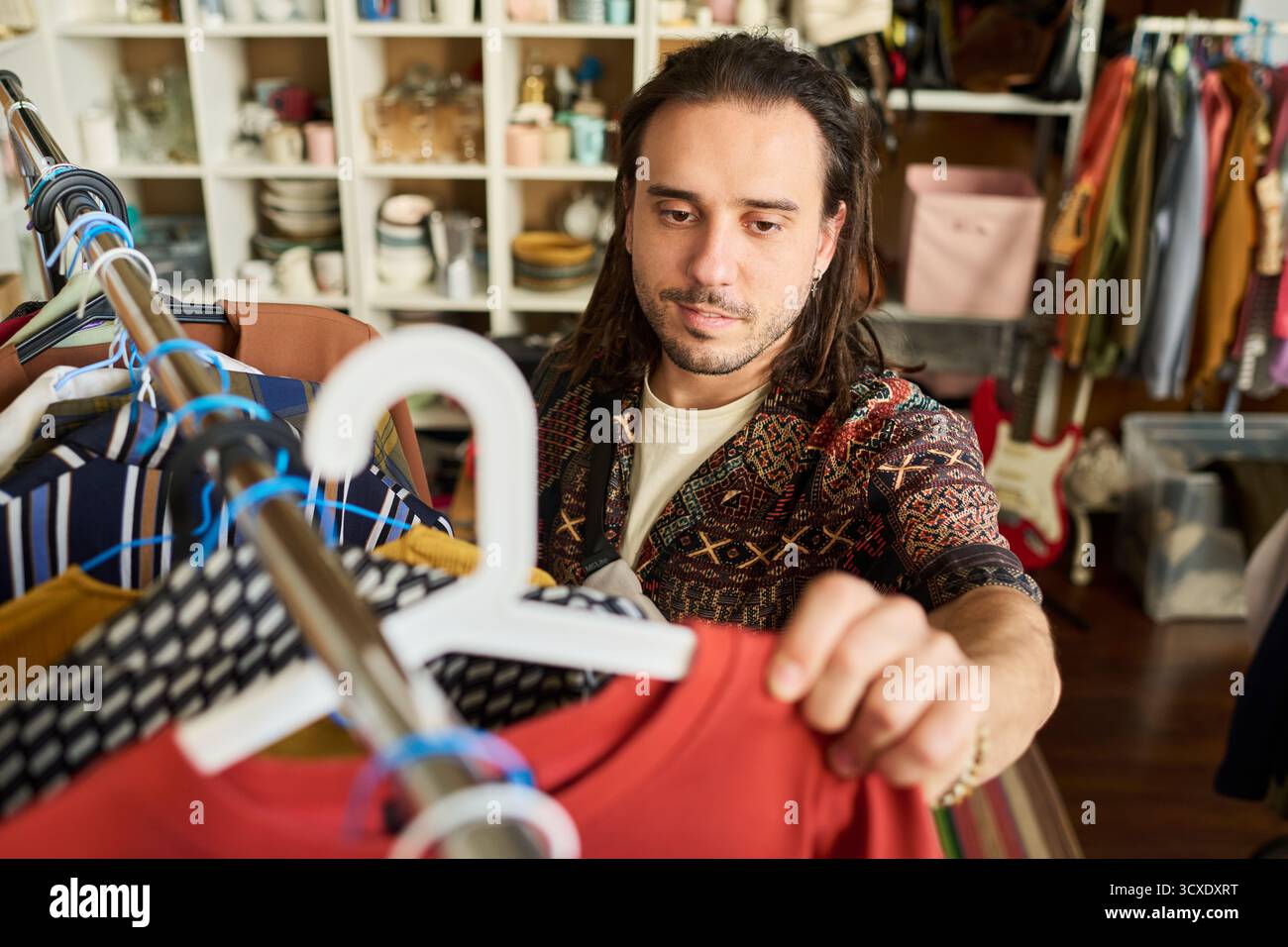 Giovane uomo adulto caucasico che sfoglia il portapacchi nel negozio di articoli di abbigliamento, selezionando capi dall'espressione mirata, circondati da articoli e accessori di seconda mano assortiti Foto Stock