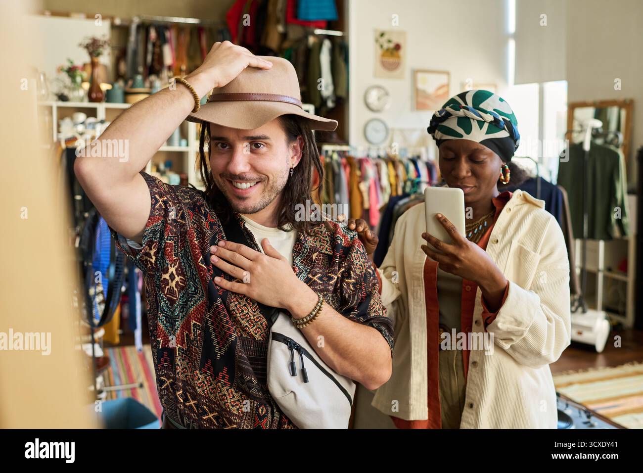 Giovane uomo adulto caucasico che prova il cappello e sorride mentre si trova in un negozio di articoli da regalo, giovane donna adulta nera in piedi nelle vicinanze che utilizza lo smartphone per scattare una foto di lui, portabiti sullo sfondo Foto Stock
